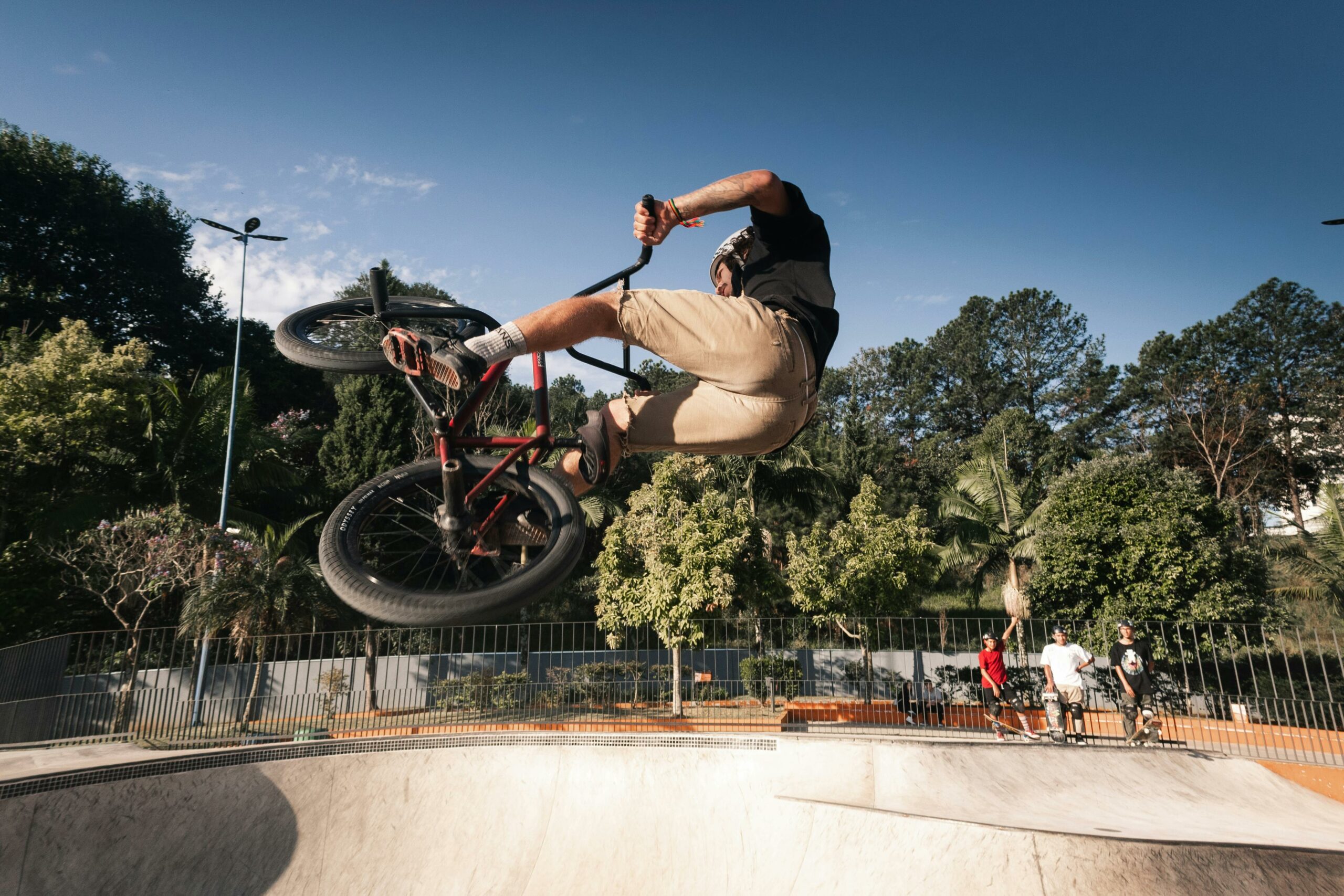 Un homme exécutant une figure en BMX dans un skatepark, suspendu en l'air au-dessus d'une rampe, avec des arbres et des spectateurs en arrière-plan.