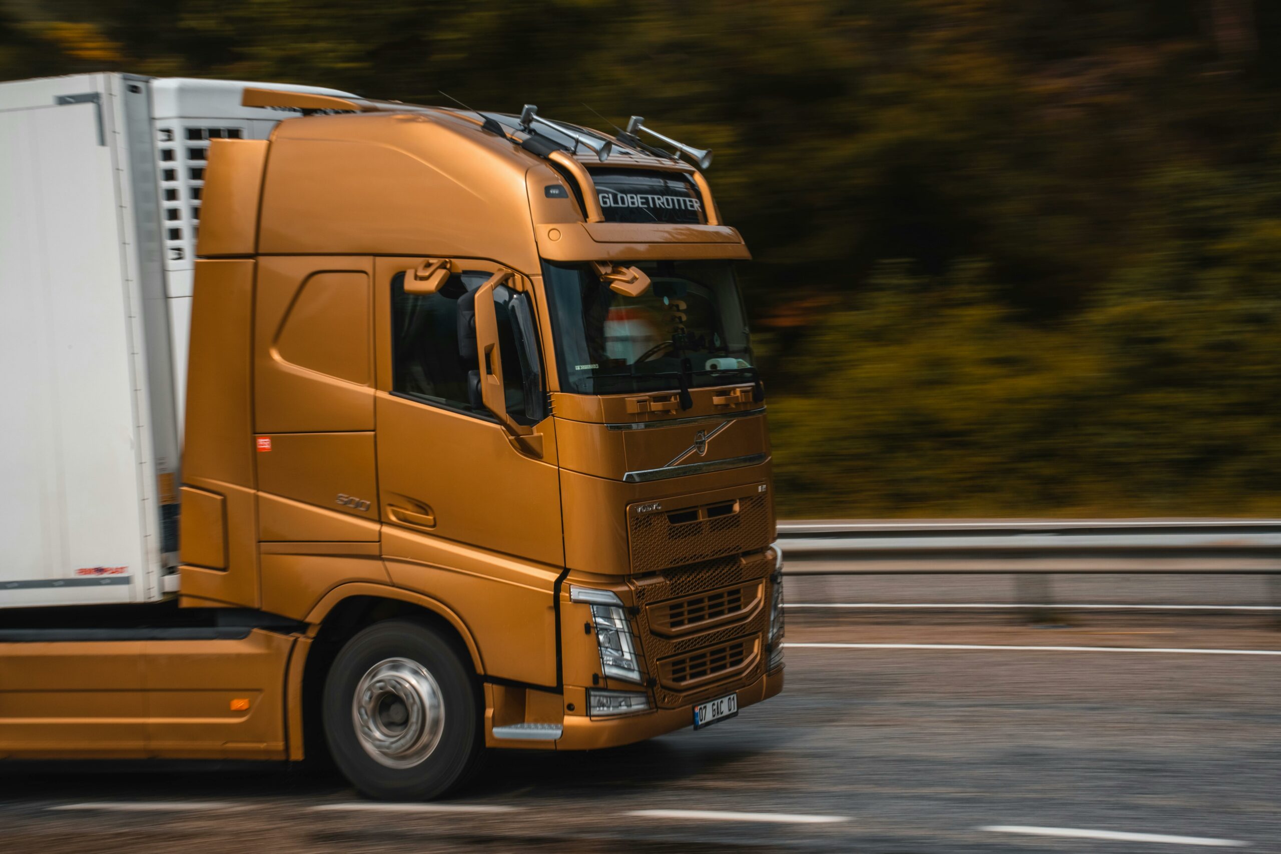 Un camion de couleur bronze transportant une remorque blanche roule rapidement sur une autoroute, avec un arrière-plan flou de végétation