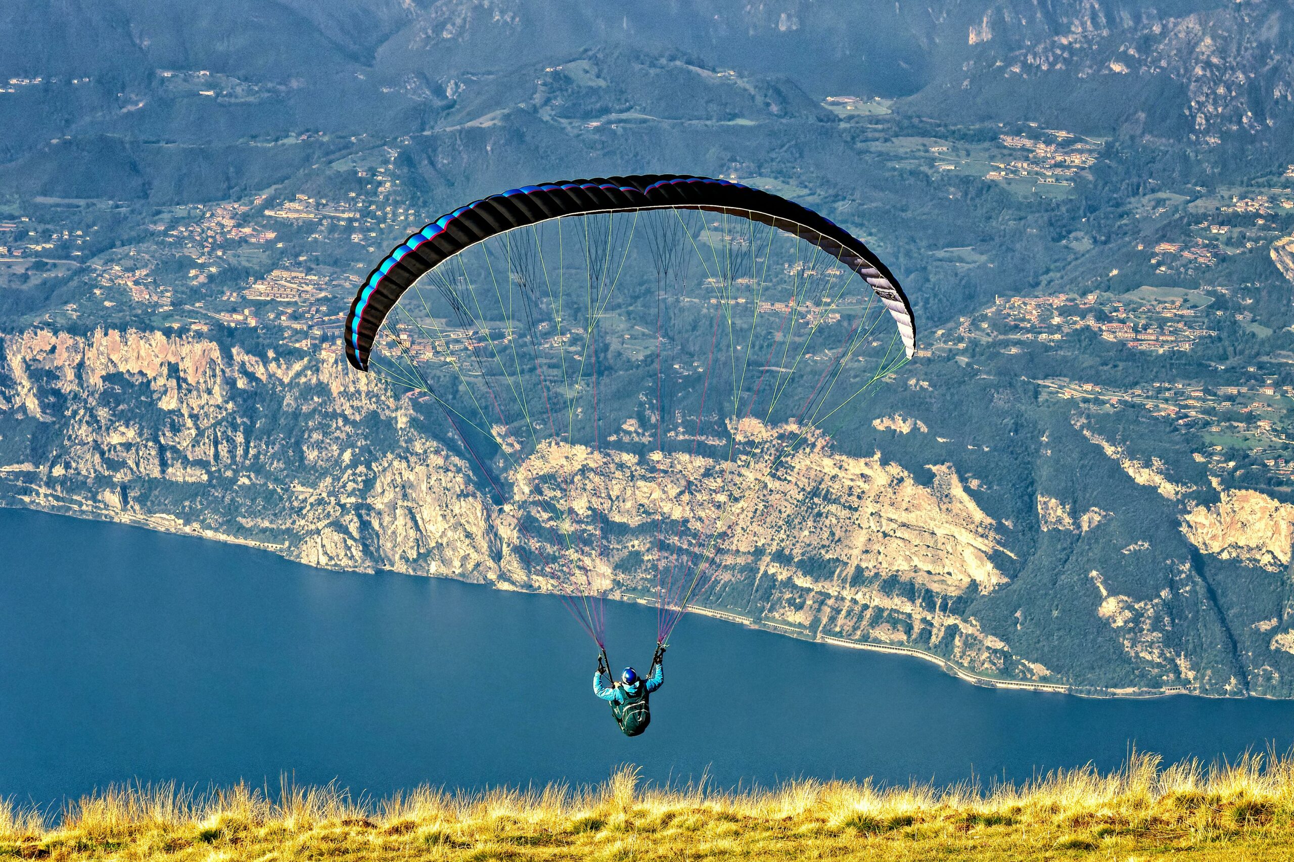 Un parapentiste survolant un lac et des montagnes rocheuses, vu de dos, avec une herbe jaunie au premier plan et un paysage vallonné en arrière-plan.
